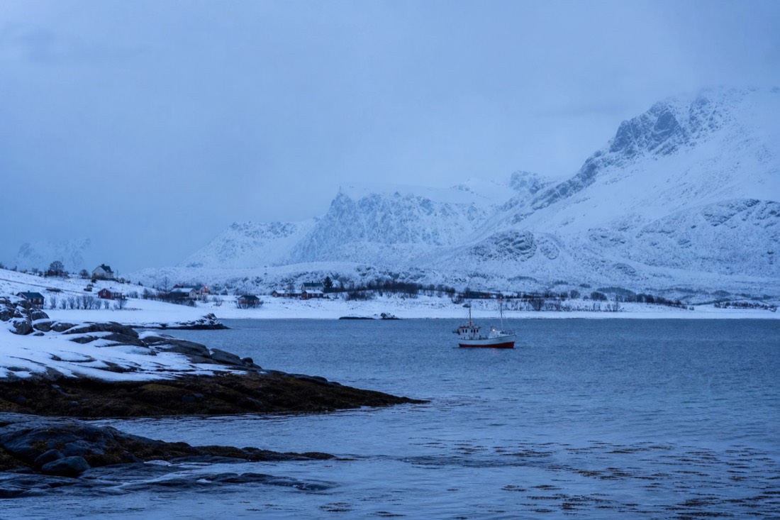 Snow-covered Arctic coastline near Tromso at blue twilight with a small fishing boat in dark water