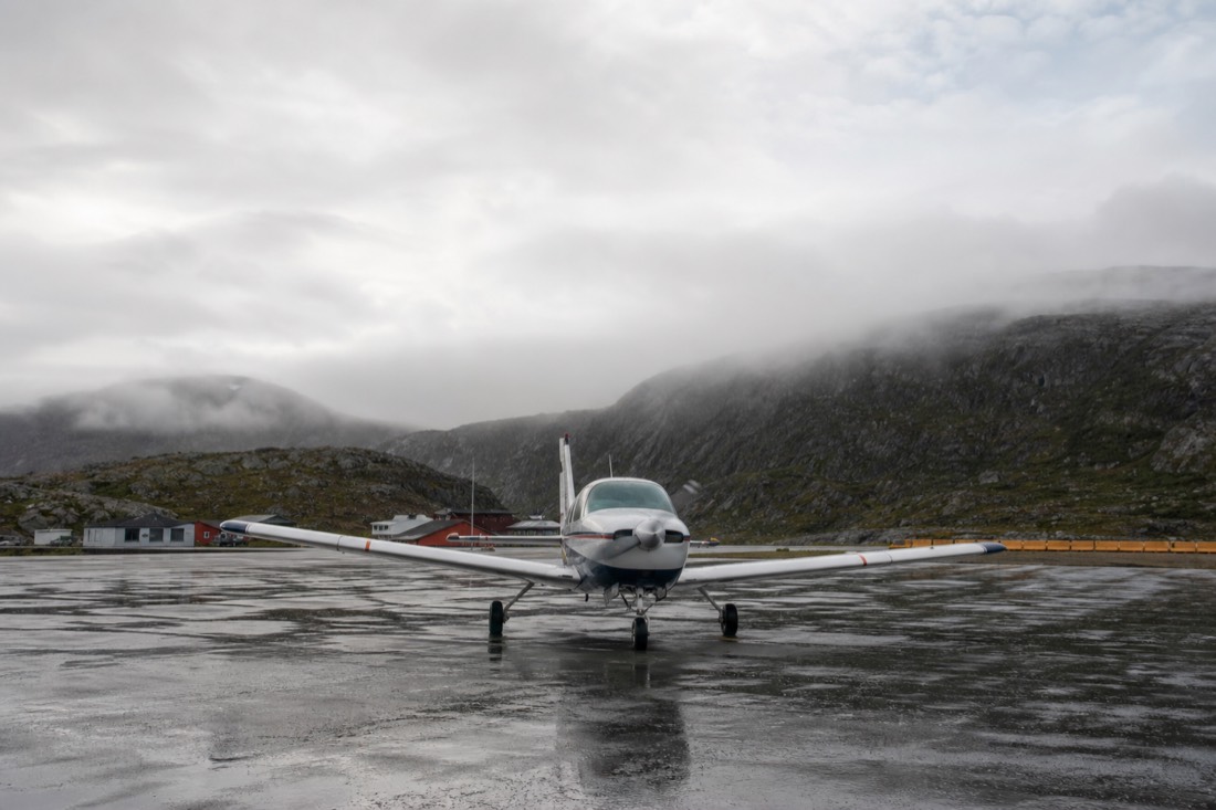 Small propeller aircraft on a wet Norwegian airport apron with low cloud over surrounding hills