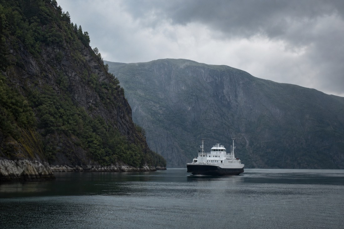 Passenger ferry crossing a still fjord between steep forested mountainsides under grey Norwegian cloud
