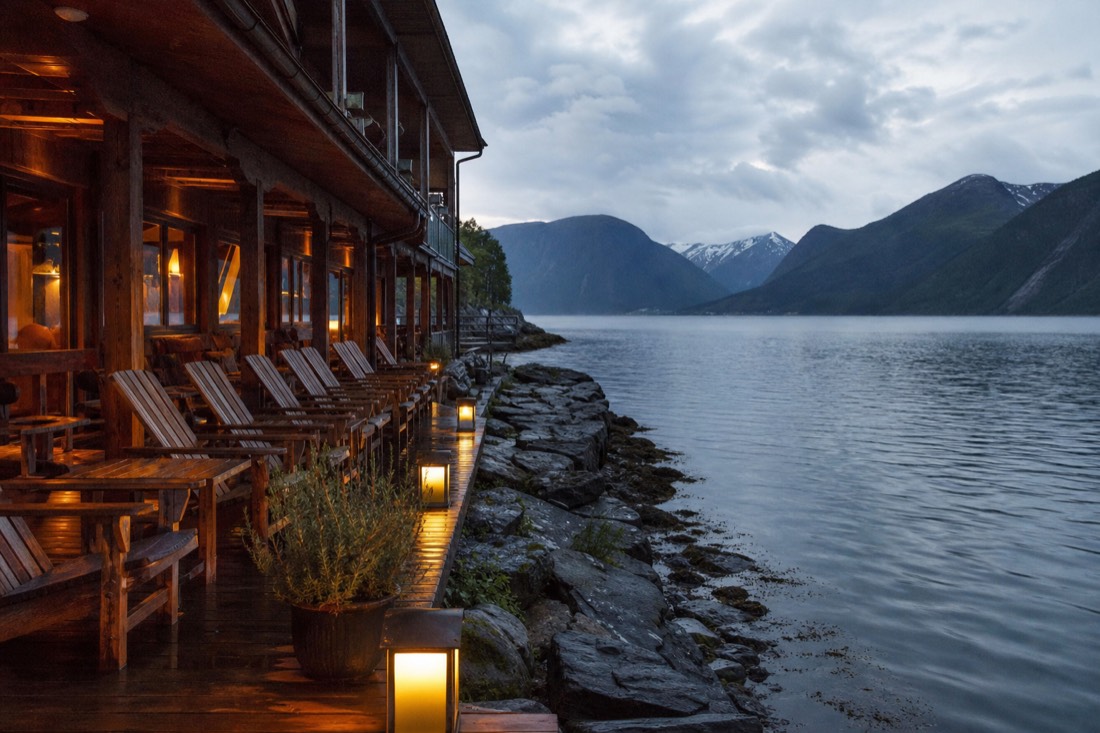 Wooden hotel terrace overlooking a fjord at dusk with warm interior lights and mountains beyond