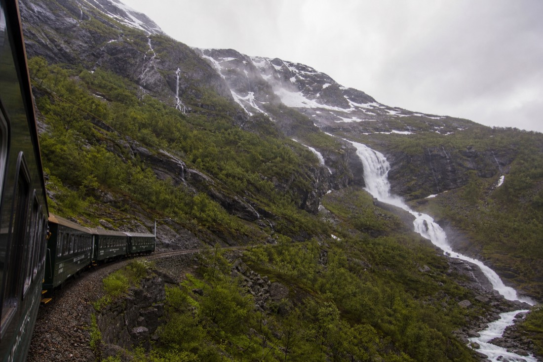Green Flåm Railway carriage descending a steep valley past a waterfall and snow patches on bare rock