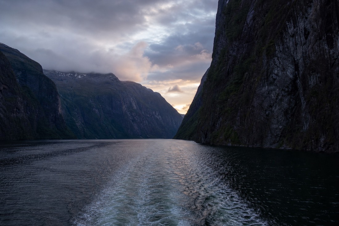 Narrow arm of Nærøyfjord at dusk with a small ferry wake cutting between sheer dark rock walls