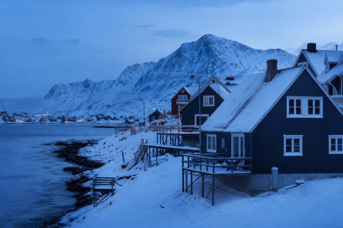 Snow-covered wooden houses in an Arctic town at blue twilight with mountains and a frozen shoreline