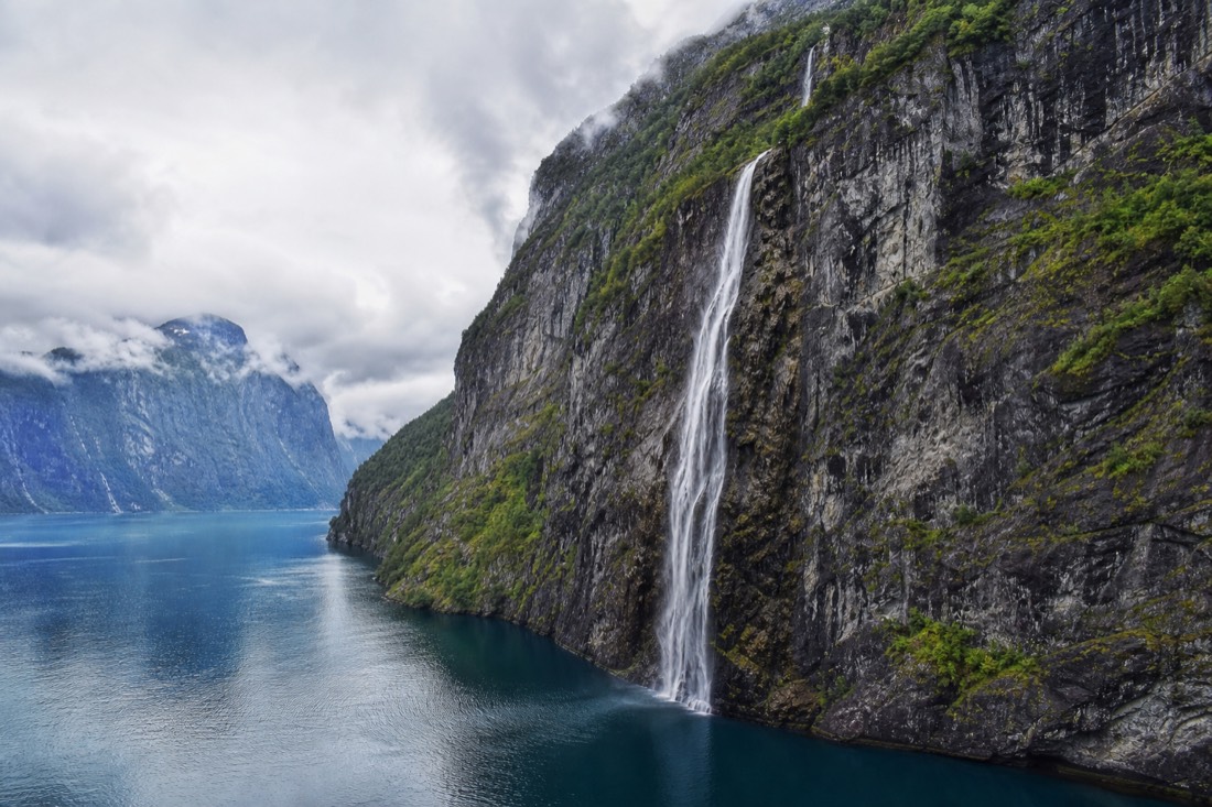 Deep blue fjord enclosed by sheer cliffs with a thin waterfall falling into the water below