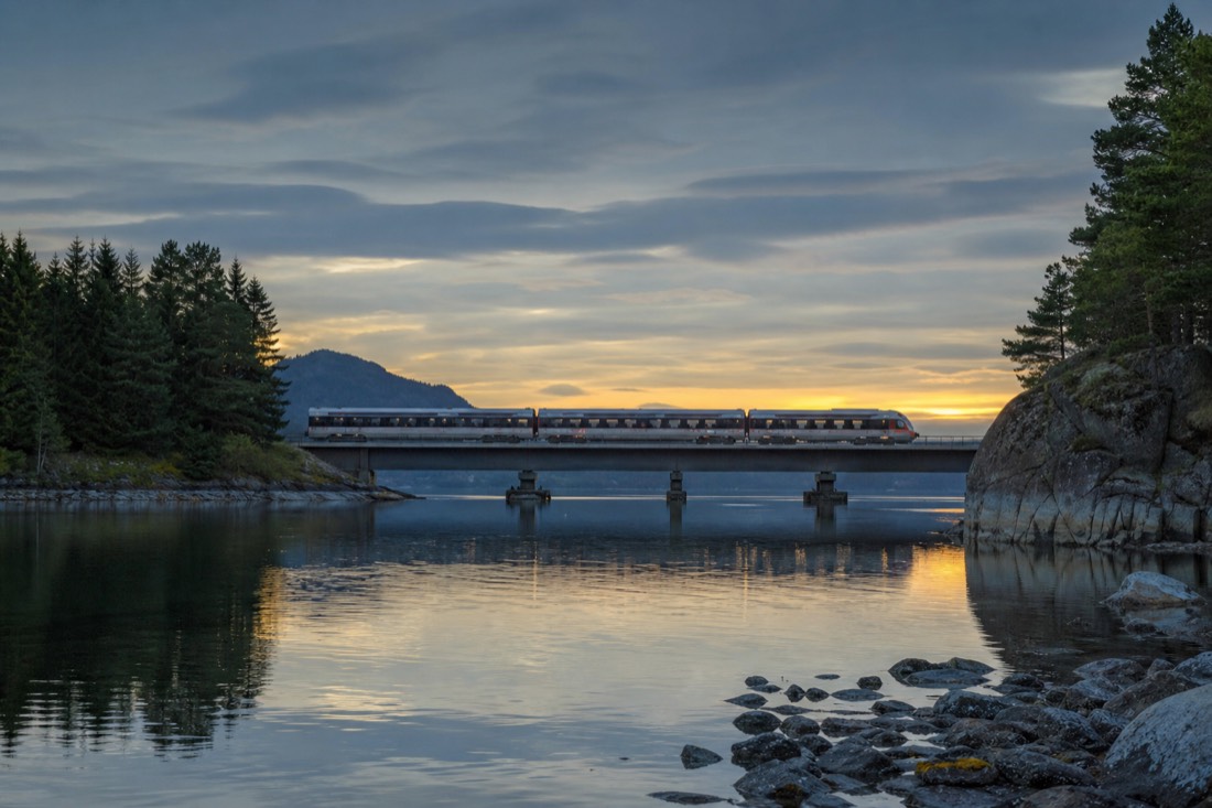 Modern train crossing a low bridge over still water between pine-covered shores at dusk