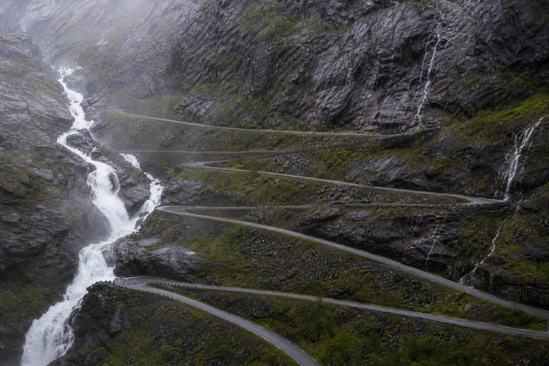 Narrow mountain road with stacked hairpin bends cut into a grey rock face beside a waterfall
