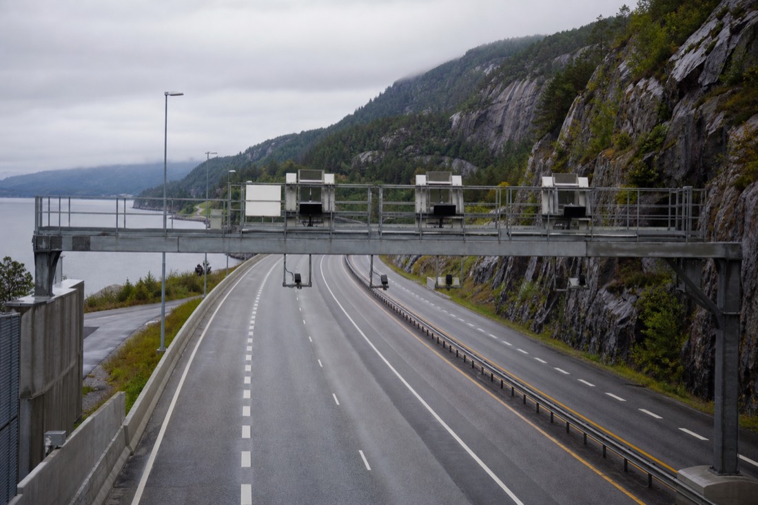 Overhead toll gantry above an empty Norwegian motorway with forested hills in the background