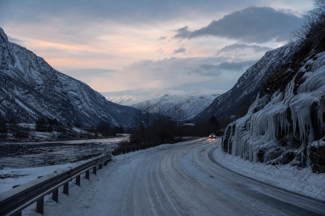 Snow-banked road through a Norwegian valley at dusk with headlights reflecting off packed ice