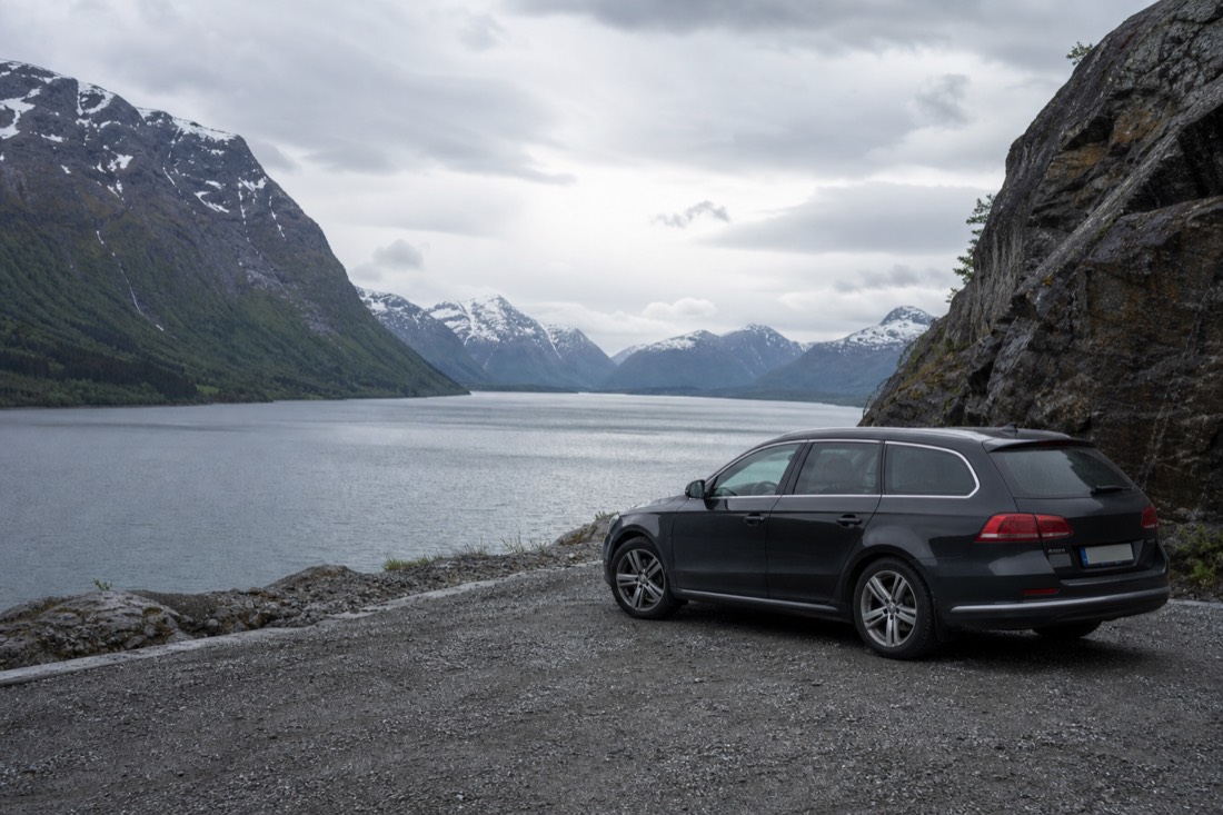 Rental car parked at a gravel viewpoint overlooking a wide fjord and distant snow-streaked peaks