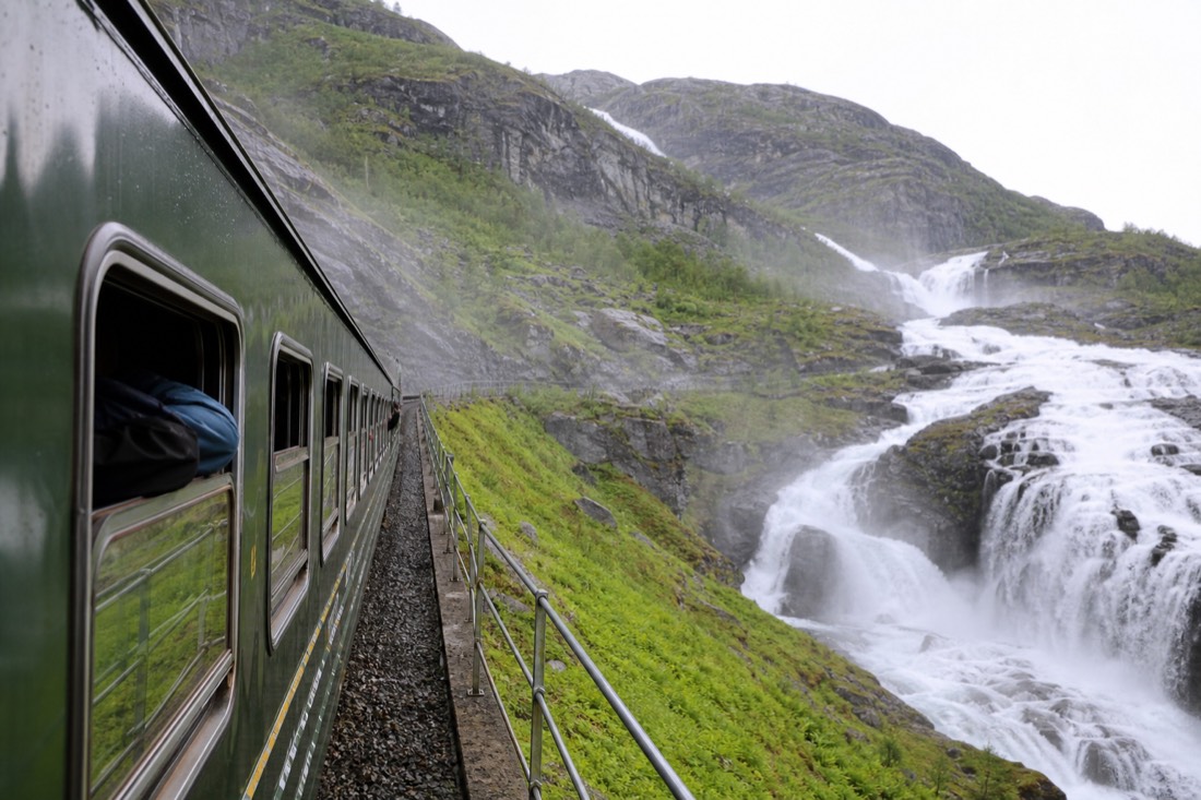 A green railway carriage stopped at a waterfall viewpoint on a steep mountain line with passengers looking out of open windows