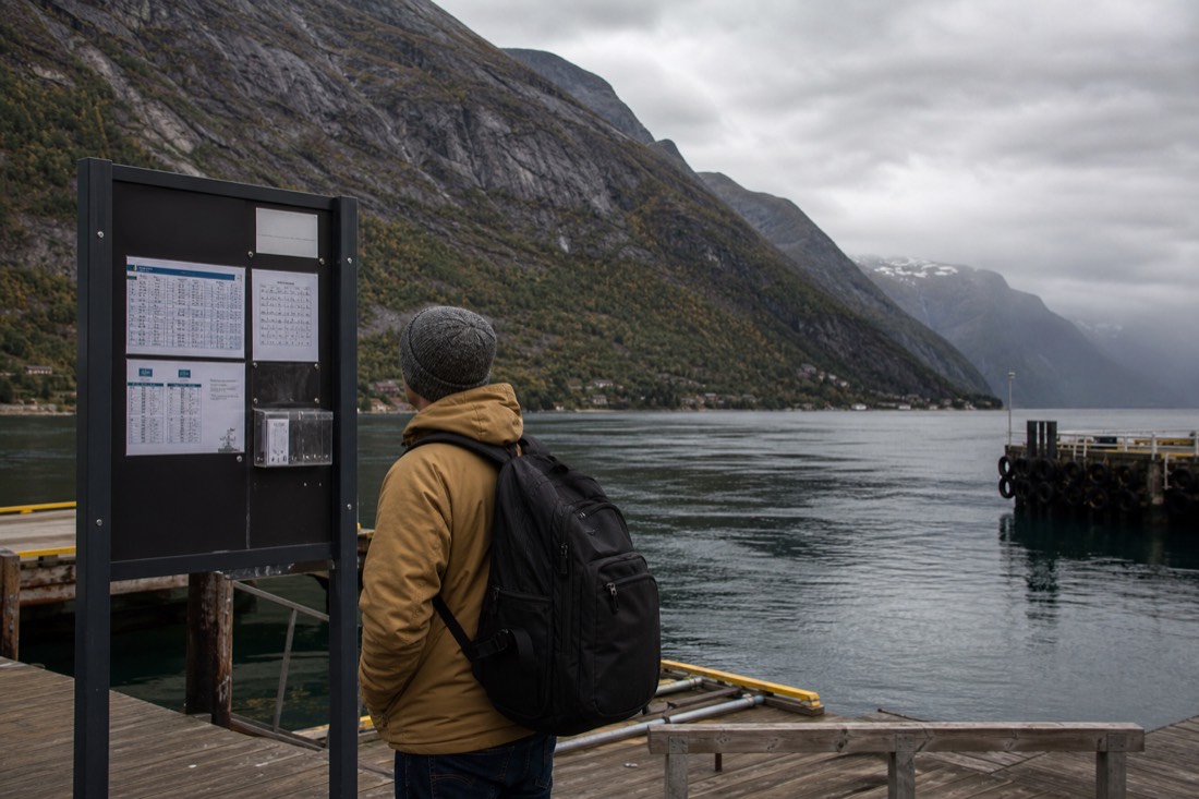 Traveller looking at a fjord ferry timetable board on a wooden quay with mountains behind