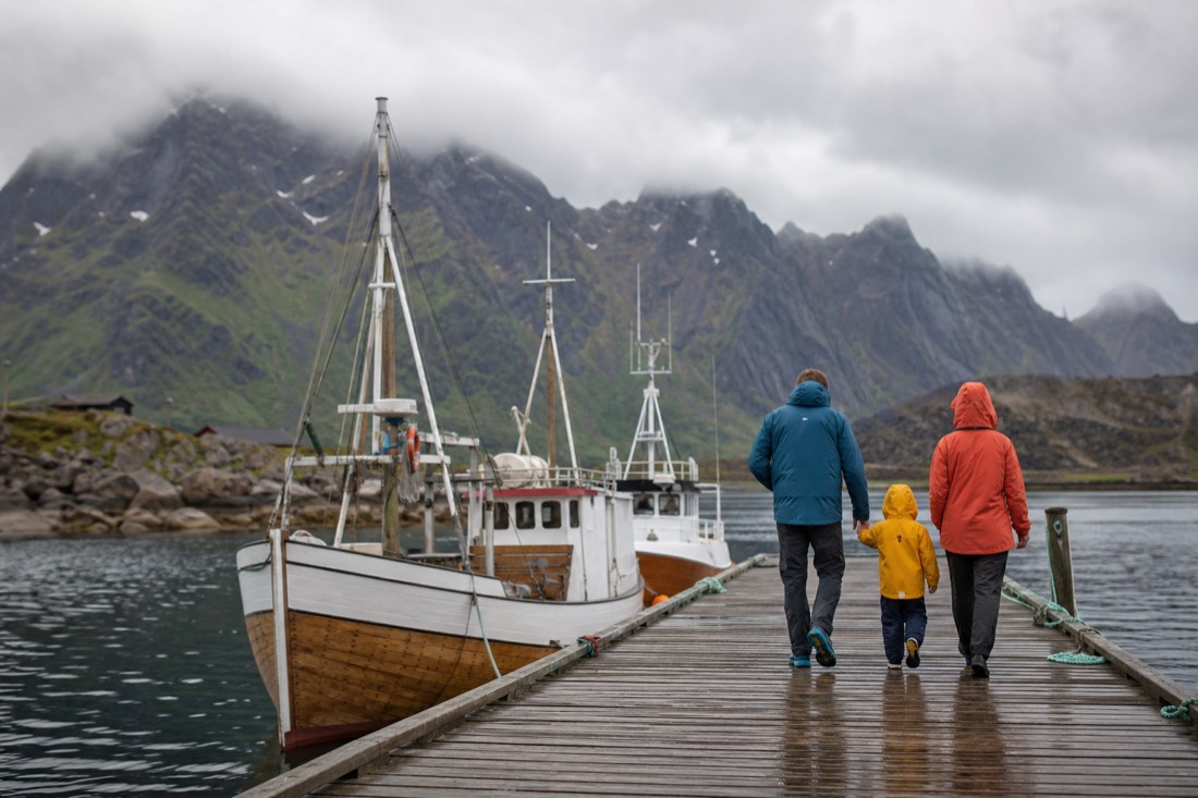 Family in rain jackets walking a wooden pier past moored fishing boats with mountains behind