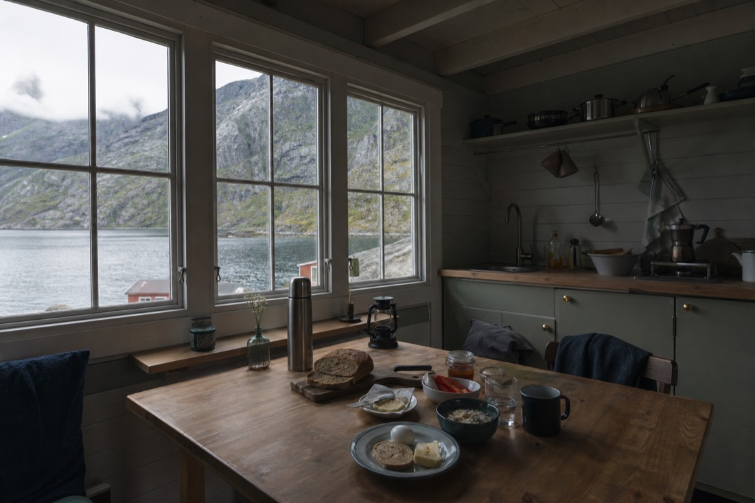 Small self-catering cabin kitchen with a wooden table set for breakfast and a fjord view through the window