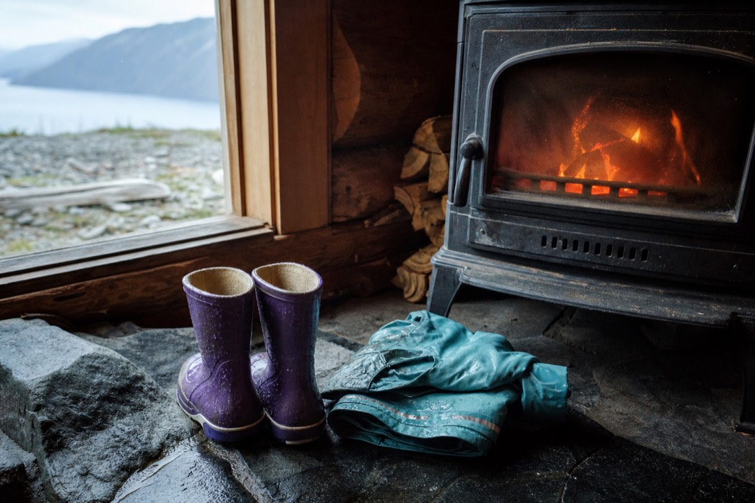 Child's rain boots and a folded waterproof jacket drying beside a wood stove in a timber cabin