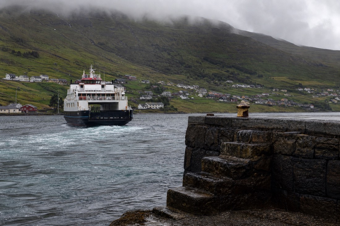 Ferry pulling away from a stone quay toward open water with steep green hills rising behind the town