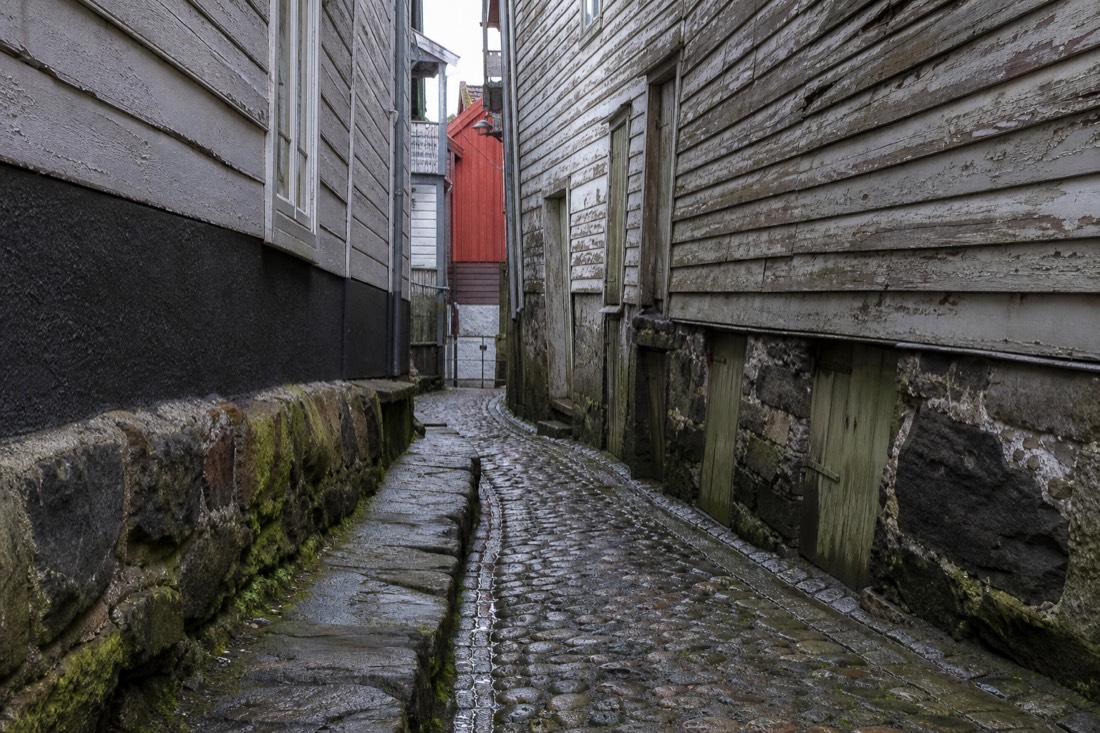 Narrow cobbled alley between leaning wooden buildings with weathered paint and a wet stone gutter