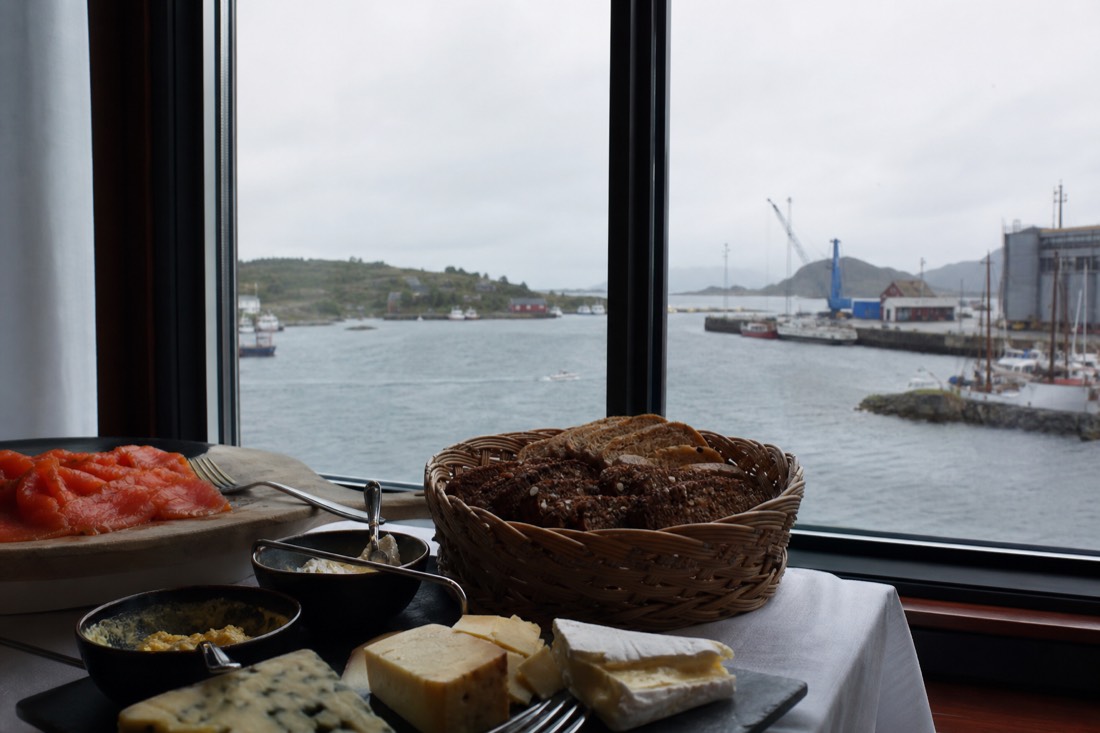 Hotel breakfast buffet of dark bread, cured fish and cheese beside a window looking onto a grey harbour