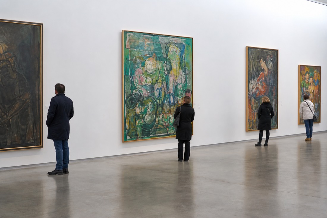 Gallery interior with high white walls, polished floor and visitors standing before large framed paintings