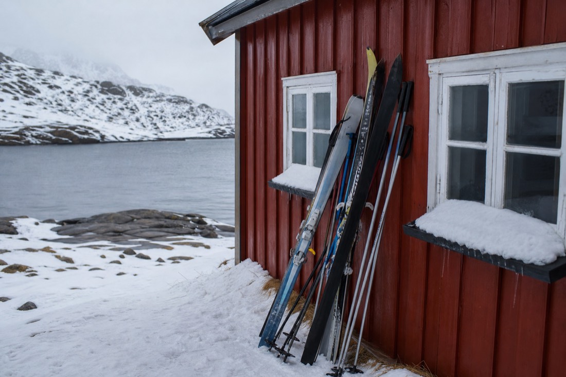 Skis and poles stacked against a red-painted mountain cabin wall with snow piled to the windowsills