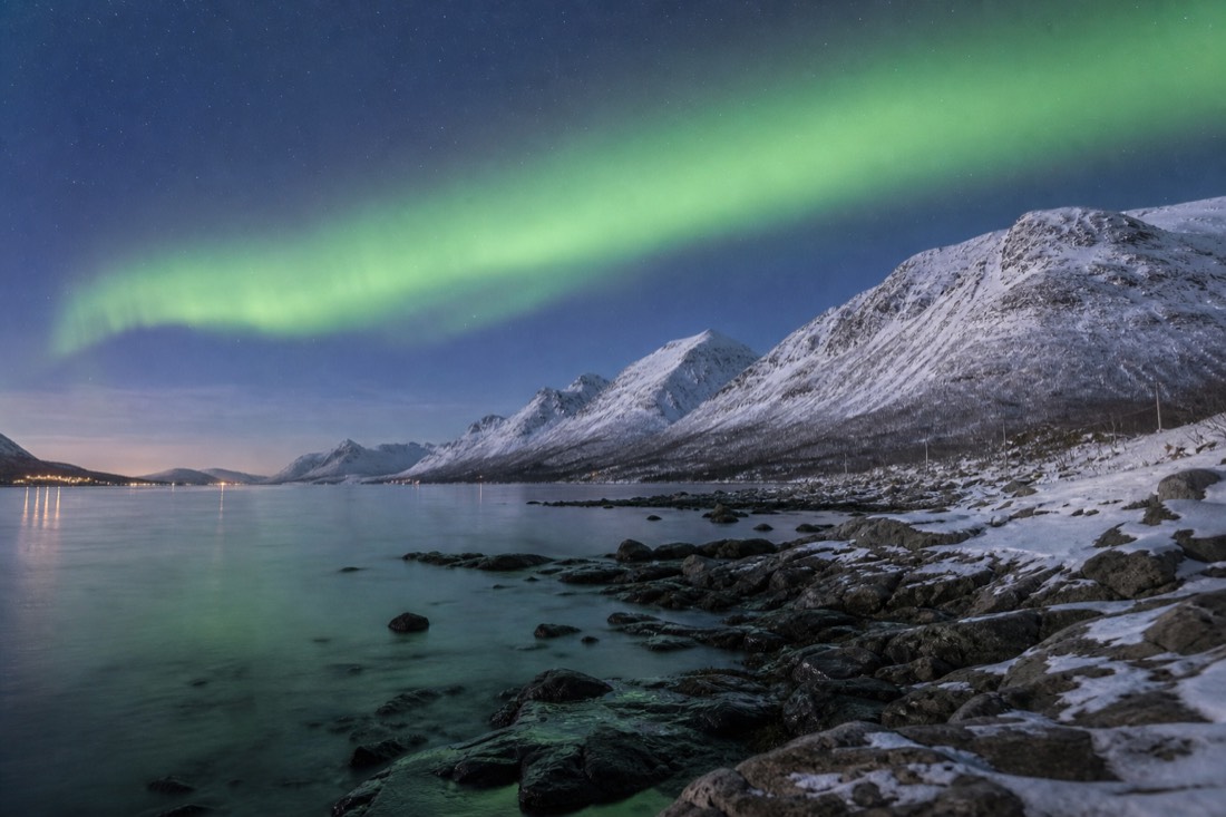 Green aurora arc over snow-covered Arctic peaks and a frozen fjord shoreline in northern Norway