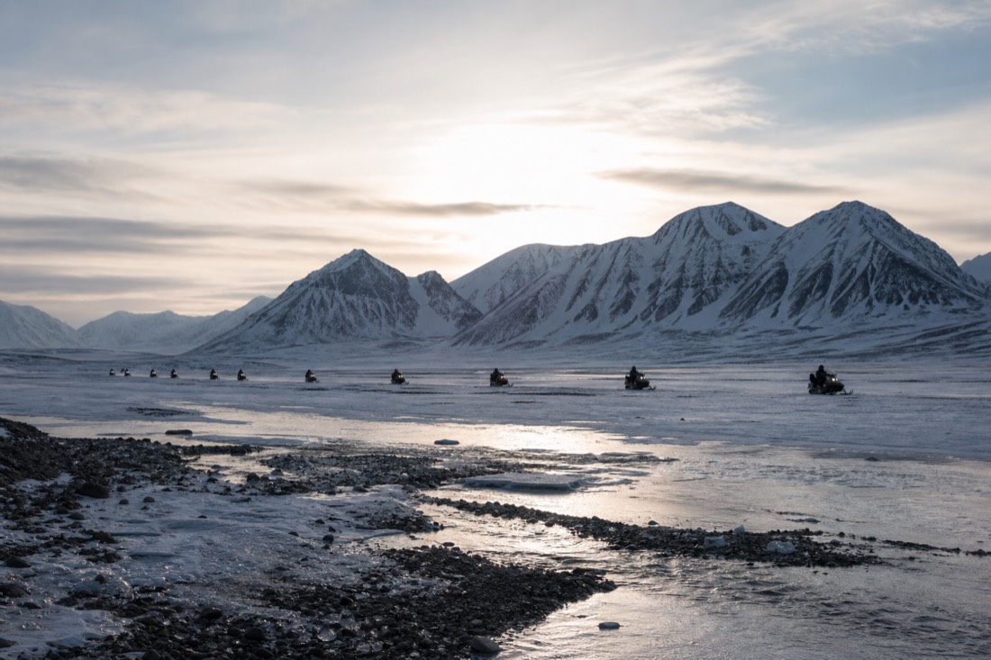 Line of snowmobiles crossing a wide frozen Svalbard valley with low sun behind sharp ridgelines