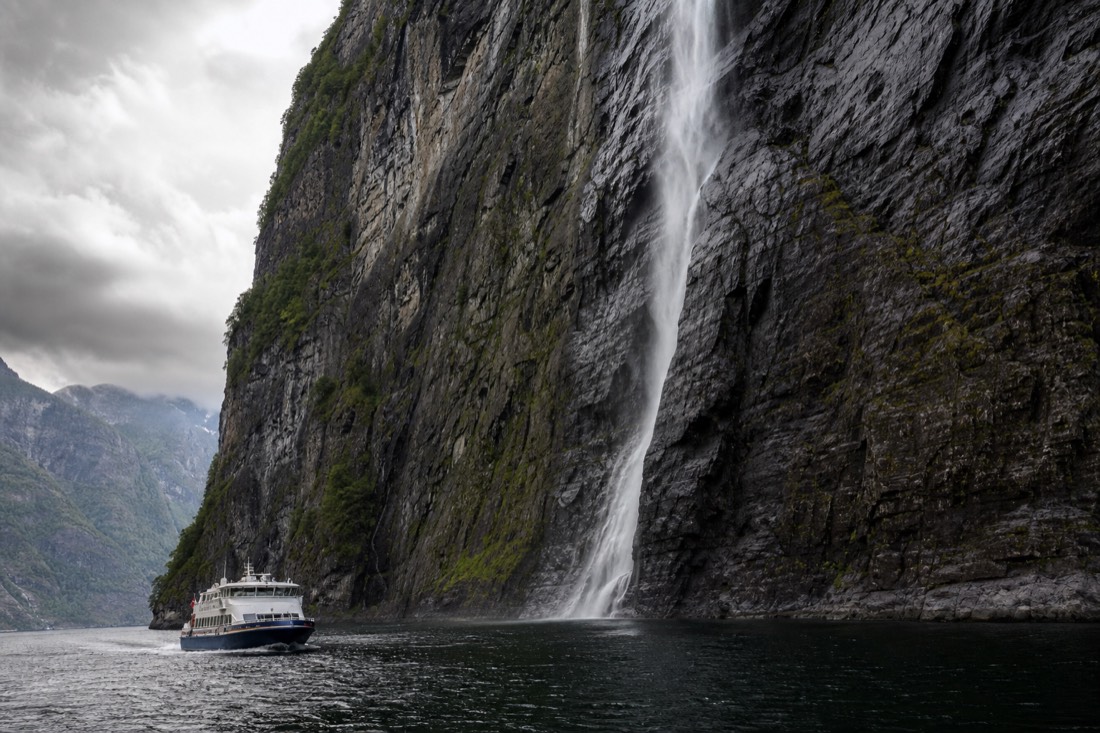 Small passenger boat close to a sheer rock wall in a narrow Norwegian fjord with a waterfall streaming down