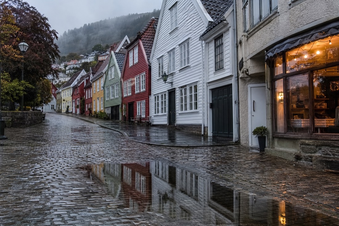 Rain-slicked cobbled street in Bergen with reflections of painted wooden houses and a lit shop window