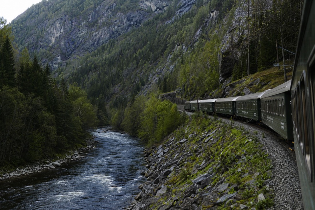 Green railway carriages curving along a steep valley wall above a river with pine forest and bare rock