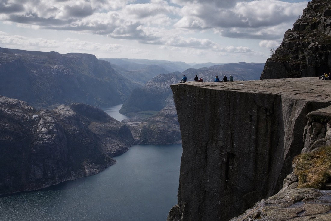 Flat cliff platform high above a long fjord with hikers sitting near the edge under scattered cloud