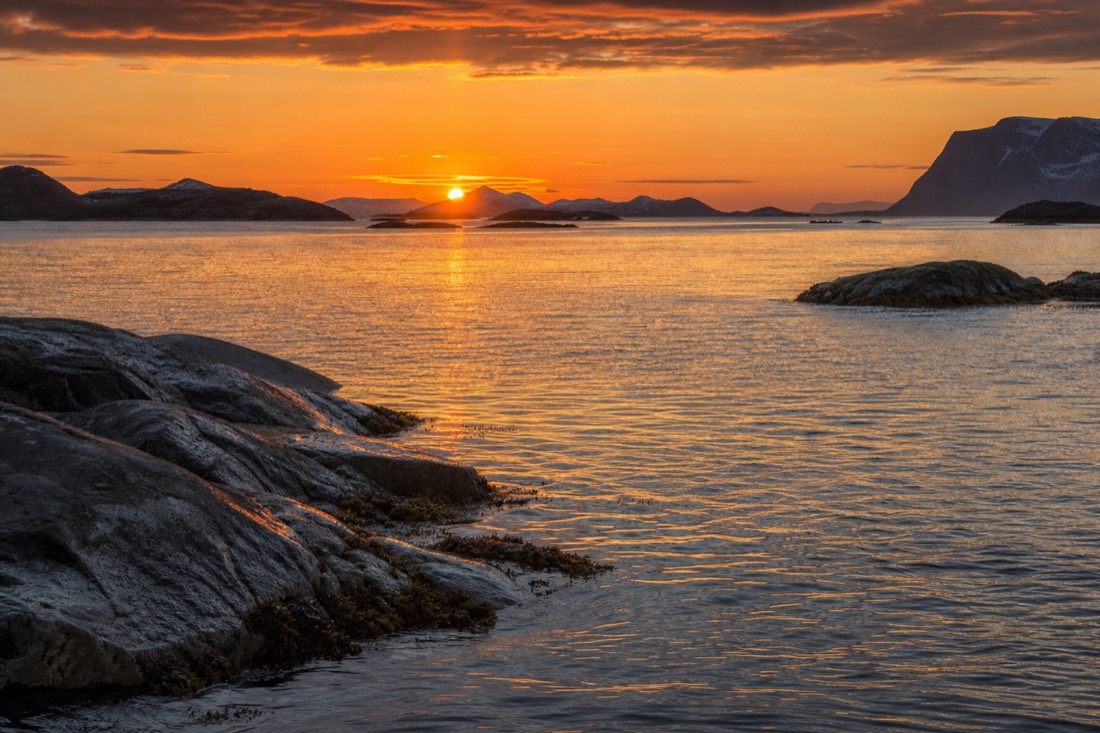 Low midnight sun burning orange over a calm Arctic sea with rocky islets in silhouette