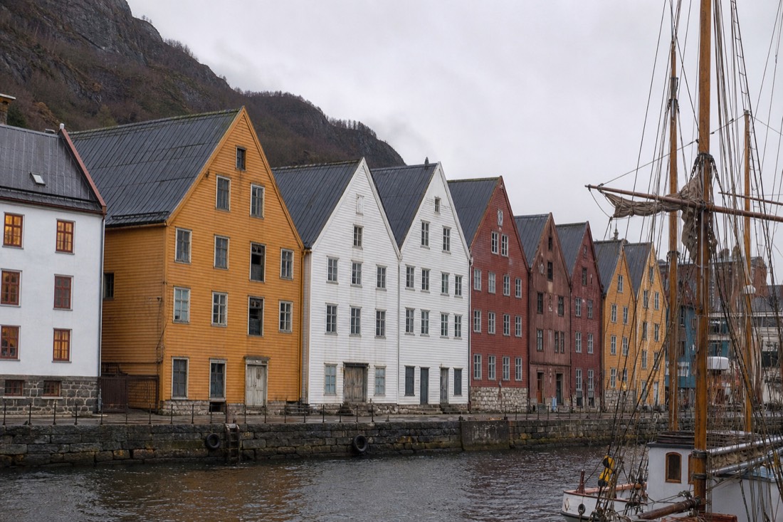 Row of tall wooden gabled warehouse fronts along a stone quay with masts and rigging in the foreground