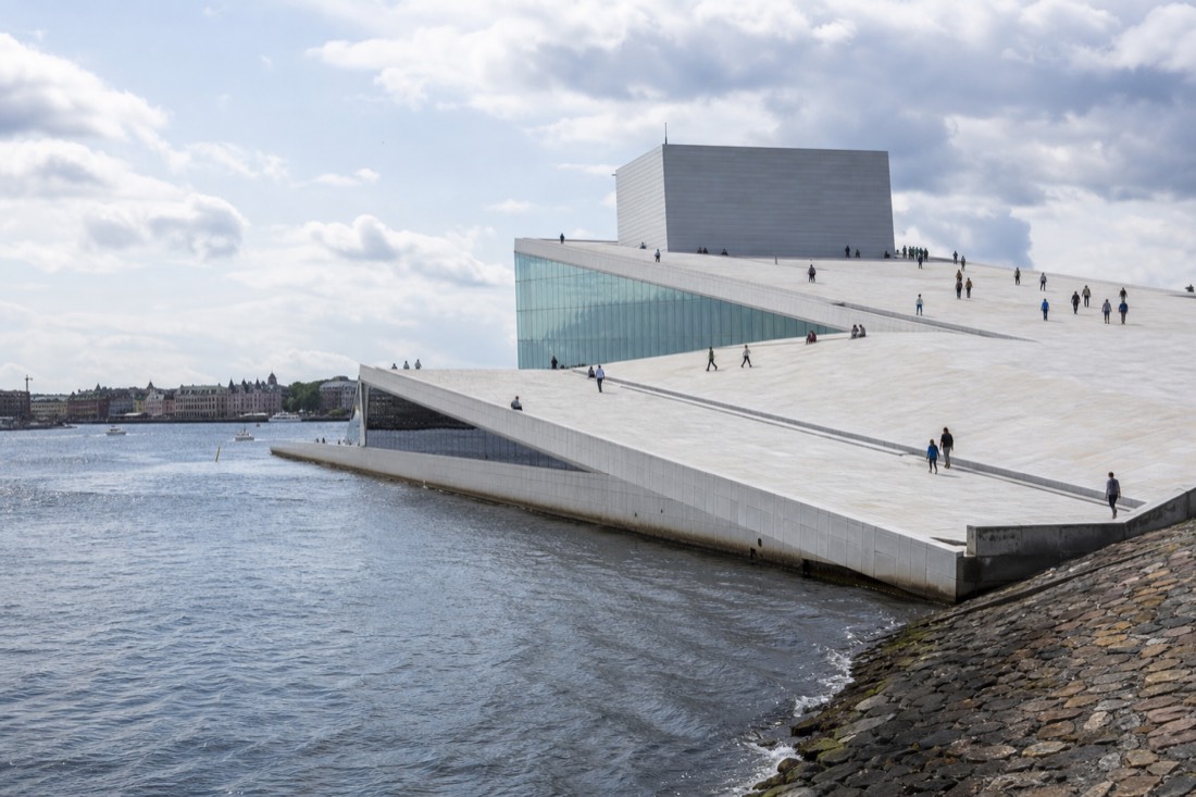 Pedestrians walking up the sloping white marble roof of a modern harbourside opera house on a bright afternoon