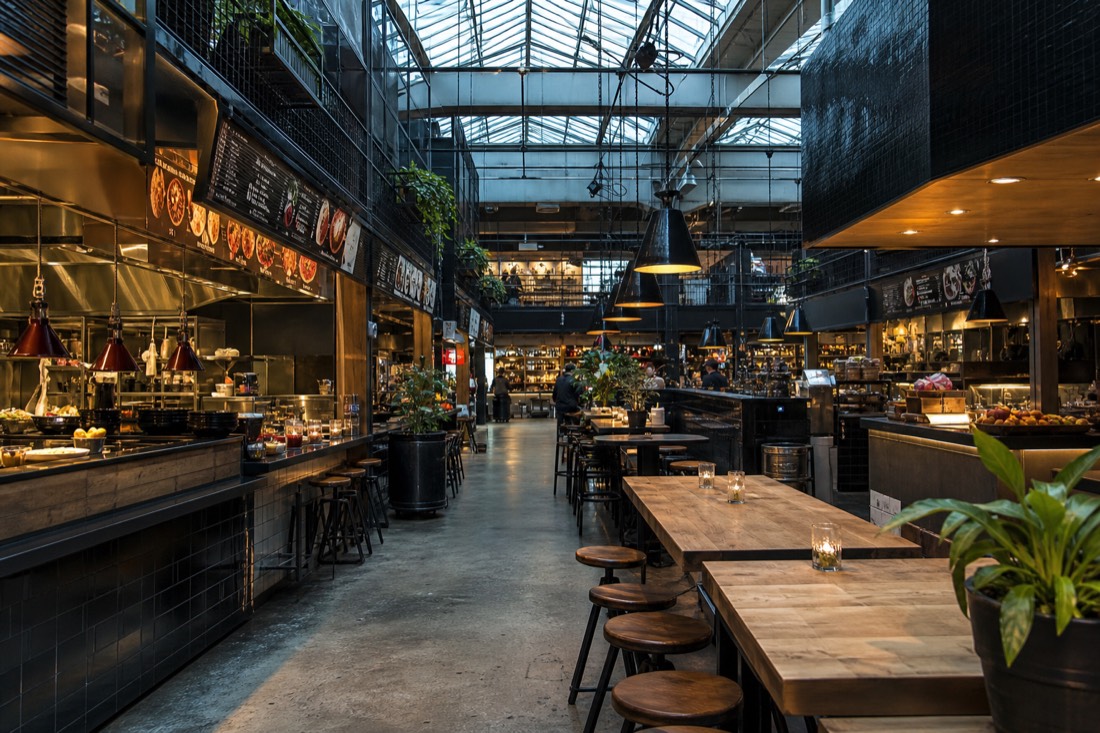 Busy indoor food hall in Oslo with counter kitchens, shared wooden tables and hanging pendant lights