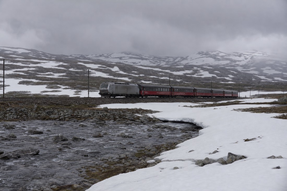 A train crossing a bare snow-streaked mountain plateau on the Oslo to Bergen railway under low grey cloud