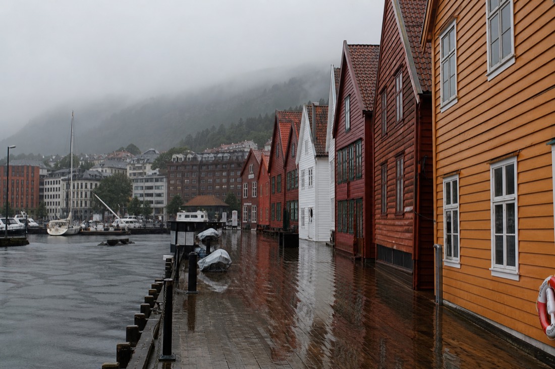 Rain-soaked wooden waterfront buildings in a Norwegian city with people walking under umbrellas